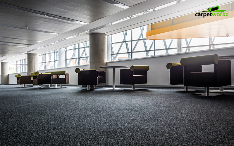 Modern office lounge area with black carpet flooring, round tables, and cushioned armchairs near large windows